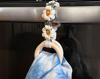 May include: A macrame towel holder featuring a white and olive green floral pattern with yellow bead centers. The holder includes a smooth, light-colored wooden ring that secures a blue and white tie-dye patterned fabric towel in place.