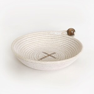 May include: A white rope bowl with a wooden bead handle. The bowl has a woven pattern and a brown X in the center.