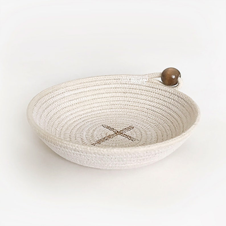 May include: A white rope bowl with a wooden bead handle. The bowl has a woven pattern and a brown X in the center.