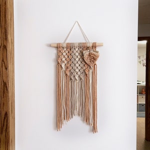 May include: A macrame wall hanging with a geometric pattern in beige and peach colors. The hanging has a wooden dowel at the top and a tassel at the bottom.