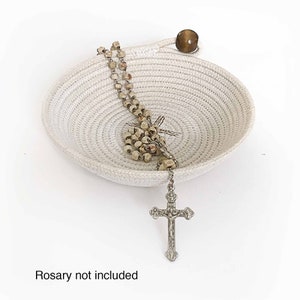 May include: A white rope bowl with a wooden handle and a silver cross and rosary beads inside. "Rosary not included" is written below the bowl.