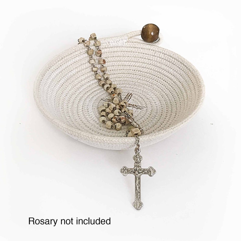 May include: A white rope bowl with a wooden handle and a silver cross and rosary beads inside. "Rosary not included" is written below the bowl.