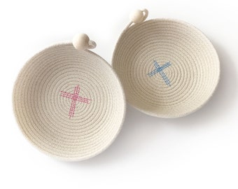 May include: Two small, round, woven baskets with wooden handles. The baskets are made of natural rope and have a cross-shaped design in the center. One basket has a pink cross and the other has a blue cross.