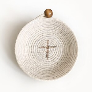 May include: A white rope bowl with a brown wooden handle and a brown cross design in the center.