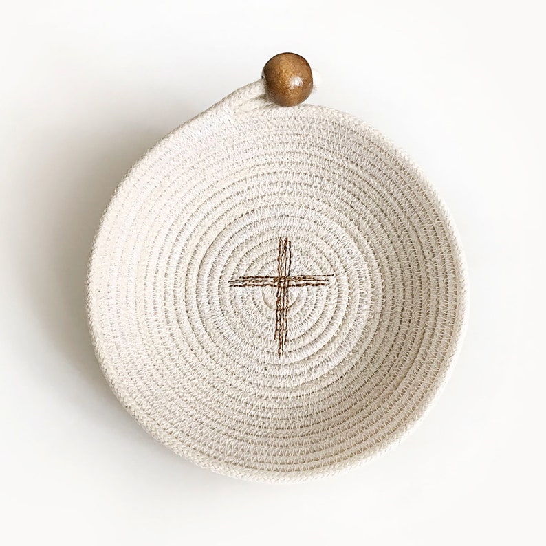 May include: A white rope bowl with a brown wooden handle and a brown cross design in the center.