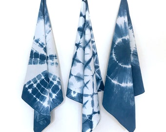 May include: Three hanging kitchen towels featuring a white and indigo blue tie-dye pattern. Each towel displays a distinct shibori-style design, including geometric grid lines, starburst motifs, and a large circular spiral pattern.