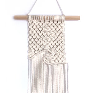 May include: Cream-colored macrame wall hanging with a wave design. The woven piece is suspended from a natural wood dowel and features knotted detailing and fringe at the bottom. A simple, elegant home decor item.