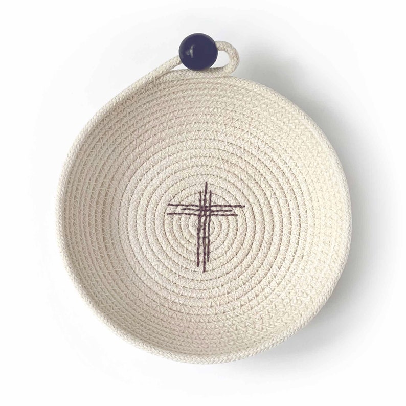 May include: A white cotton rope bowl with a dark blue knob handle and a brown embroidered cross in the center.