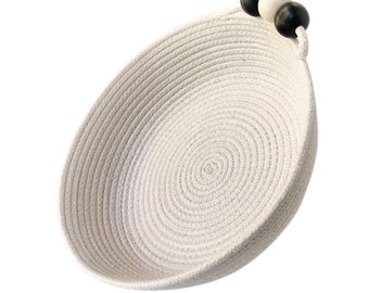 May include: A white rope bowl with a black and white beaded handle. The bowl is round and has a slight curve to it. The beads are made of wood and are strung together with a white cord.