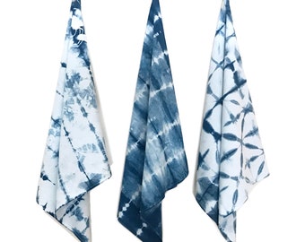 May include: Three hanging kitchen towels featuring distinct blue and white tie-dye patterns. Each towel displays a unique indigo-colored design, including vertical stripes, diagonal bands, and a geometric grid motif on a white fabric base.
