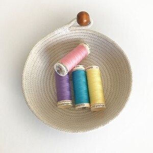 May include: A white rope bowl with a wooden knob handle holds four spools of thread. The spools are pink, purple, blue, and yellow. The spools have German text on them.