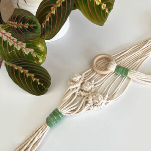 May include: A cream-colored macrame plant hanger with a wooden ring and green accents. The plant hanger is made of woven cotton rope and features decorative knots. A Maranta plant with green leaves and red veins is visible in the background.