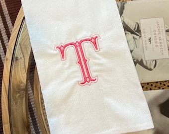 Può includere: Asciugamano bianco con la lettera "T" ricamata in rosa. L'asciugamano è piegato e appoggiato su un tavolo con piano in vetro. Sullo sfondo è visibile un libro con il testo "Jackie Kennedy Onassis".