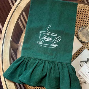 May include: A dark green kitchen towel with a white embroidered coffee cup design and the word "Ralph's". The bottom edge features a ruffled trim. The towel is displayed on a round table.