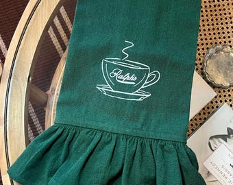 Può includere: Un canovaccio da cucina verde scuro con un disegno di tazza da caffè ricamata in bianco e la scritta "Ralph's". Il bordo inferiore presenta un bordo a balze. L'asciugamano è esposto su un tavolo rotondo.