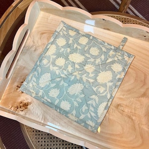 May include: A square, quilted potholder with a floral pattern in light blue and cream, resting on a wooden serving tray. The tray has a scalloped edge and a light finish. The potholder has a small loop for hanging.