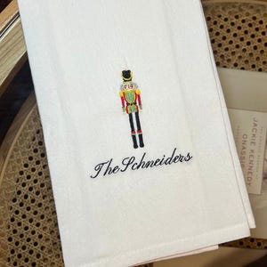 May include: White embroidered towel featuring a colorful nutcracker design and the name "The Schneiders" in elegant script. The nutcracker is detailed with red, green, and gold accents, adding a festive touch to the towel.