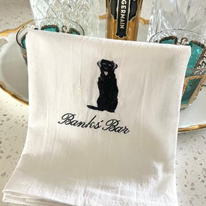 May include: White cotton bar towel with a black embroidered Labrador Retriever design holding a champagne glass. The text "Banks' Bar" is embroidered below the dog. The towel is folded and placed on a white surface.