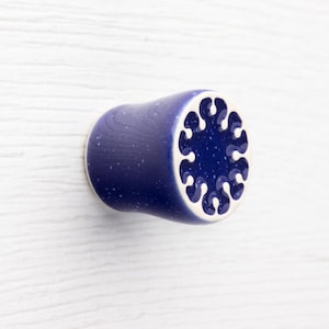 Op de afbeelding: Een keramische kastknop met een cilindrische vorm. De knop is diepblauw met witte spikkels en een decoratieve witte rand. De voorkant van de knop heeft een donkerblauw, gestileerd bloemmotief.