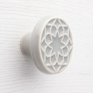 May include: A light grey ceramic cabinet knob featuring a star and geometric design. The knob is round with a stem for fixing. The pattern is light grey, and the knob is set against a white wooden surface.