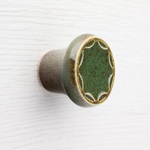 May include: A ceramic cabinet knob with a green and brown finish. The knob has a circular face with a green centre and a decorative white pattern. The knob is mounted on a white wooden surface.