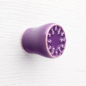 May include: A purple ceramic ear plug with a decorative design on the end. The plug is cylindrical with a flared end featuring a raised, swirling pattern. The colour is a deep, glossy purple.