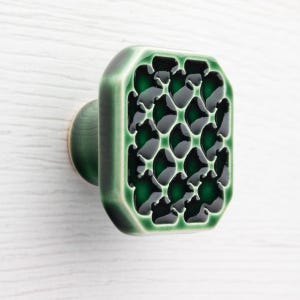 Könnte beinhalten: Ein grüner Keramik-Schrankknauf mit rechteckiger Front mit einem dunkelgrünen, gemusterten Design. Der Knauf hat abgerundete Kanten und eine kreisförmige Basis, montiert auf einer weißen Holzoberfläche.