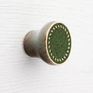 Könnte beinhalten: Ein Keramik-Schrankknopf mit abgerundeter Form. Der Knopf hat eine dunkelgrüne Mitte mit strukturierter Optik, umgeben von einem Ring aus kleinen weißen Punkten. Der Körper des Knopfes hat eine gedämpfte grün-braune Farbe.
