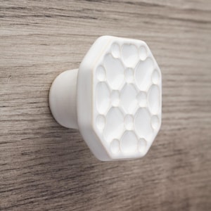 Puede incluir: Pomo de armario de cerámica blanca con un patrón geométrico texturizado. El pomo tiene una cara octogonal con una serie de formas redondeadas en relieve. El pomo está montado sobre una superficie de madera, ofreciendo una estética limpia y moderna.