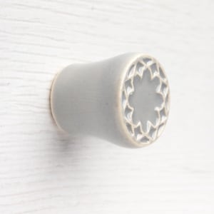 May include: A light gray ceramic cabinet knob with a smooth, flared cylindrical body and a flat circular face featuring a recessed, star-shaped geometric pattern with intricate cutouts around the center, finished with a subtle glossy glaze.