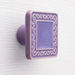 May include: A square, purple ceramic cabinet knob with a decorative frame and a round base. The centre of the knob is a lighter shade of purple, and the frame has a cream-coloured scroll design.