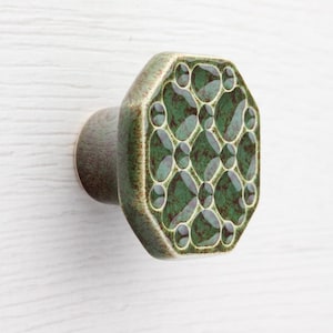 May include: A green ceramic cabinet knob with a geometric pattern. The knob has an octagonal face with a dark green and light green design. The base of the knob is a muted green color.