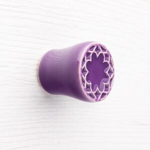 May include: A purple ceramic ear plug with a starburst design on the end. The plug is a solid, glossy purple with a hollowed-out center featuring a white star pattern. The design is symmetrical and detailed.