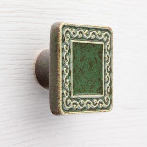 May include: A square cabinet knob with a green centre and decorative border. The knob has a bronze-coloured base and a rectangular face with a textured green surface and a white and green scroll design border.