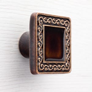 Könnte beinhalten: Ein dekorativer Schrankknopf mit rechteckiger Front und rundem Griff. Die Front ist dunkelbraun mit einem helleren braunen Zierrand. Der Griff ist dunkelbraun. Der Knopf ist auf einer weißen Oberfläche montiert.