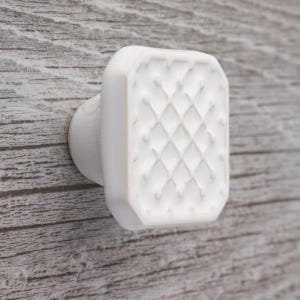 Könnte beinhalten: Weißer, rechteckiger Schrankknauf mit einem Rautenmuster auf der Vorderseite. Der Knauf ist auf einer Holzoberfläche montiert. Das Design des Knaufs verleiht jedem Schrank oder jeder Schublade einen Hauch von Eleganz.