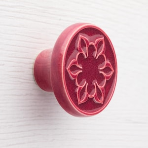 Può includere: Un pomello per armadio in ceramica rossa con un design floreale. Il pomello ha una forma arrotondata con una faccia circolare con un motivo floreale in rilievo. Il colore è un rosso intenso e ricco, e il pomello è montato su una base di legno.