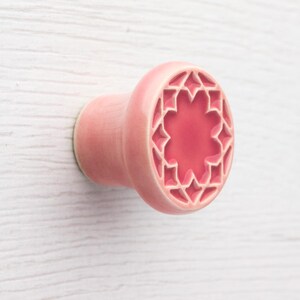 Könnte beinhalten: Ein rosa Keramik-Schrankknopf mit Sternmuster. Der Knopf hat eine abgerundete Form mit zylindrischem Sockel und einer dekorativen Oberfläche mit geometrischem Muster. Die Farbe ist ein weiches, gedämpftes Rosa.