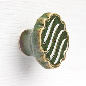 May include: A ceramic cabinet knob with a scalloped edge and a glossy finish. The surface features a mottled green and brown glaze with three distinct, diagonal white stripes across the centre. The knob has a rounded, tapered base.
