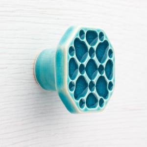 Puede incluir: Pomo de armario de cerámica turquesa con un diseño único. El pomo tiene una cara octogonal con un patrón de formas interconectadas. Montado sobre una superficie de madera blanca. Mide aproximadamente 5 cm de largo.