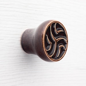 Könnte beinhalten: Dunkelbrauner Holzschrankknauf mit einem dekorativen, wirbelnden Design auf der Vorderseite. Das Design besteht aus einer Reihe miteinander verbundener, geschwungener Linien in einem dunkleren Braunton vor einem helleren braunen Hintergrund.