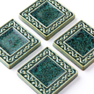 May include: Four square ceramic coasters with a diamond shape and a teal green mottled finish. Each coaster has a raised border with a cream-colored, scroll-like design. The coasters are arranged on a white surface.