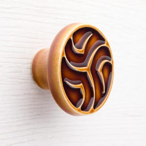 Puede incluir: Pomo decorativo para armario con base de madera marrón claro y cara redonda de cerámica. La cara presenta un diseño abstracto marrón oscuro con acabado brillante. El pomo está montado sobre una superficie de madera blanca.
