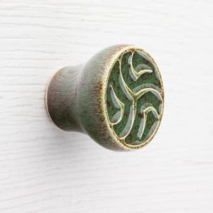 Könnte beinhalten: Keramik-Schrankknopf in Grün und Braun. Der Knopf hat eine runde, grüne, geschnitzte Musterfläche. Montiert auf einer weißen Oberfläche, ideal für Möbeldekoration und Heimwerkerprojekte.