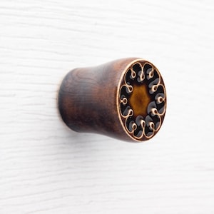 May include: A dark brown wooden ear plug with a decorative face. The face features a gold-colored border with a repeating black, swirling design around a central, amber-colored circle. The plug is cylindrical with a flared end.