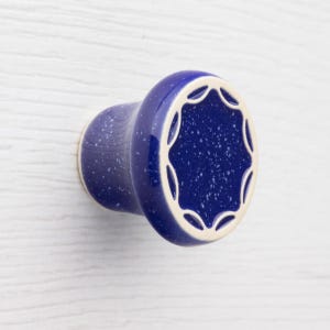 Op de afbeelding: Een blauwe keramische kastknop met een gespikkelde afwerking. De knop heeft een afgeronde vorm met een decoratieve geschulpte rand in crème. Deze hardware is ontworpen voor laden en kasten.