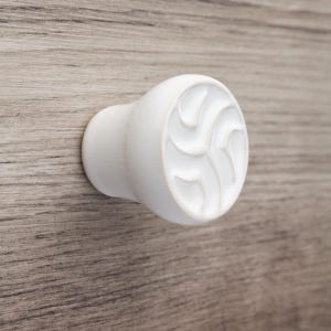 Può includere: Un pomello per armadio rotondo bianco con un motivo a spirale decorativo. Il pomello è realizzato in legno ed è montato su una superficie in legno con un motivo a venature.