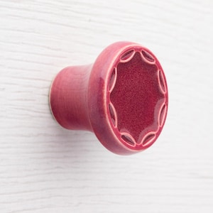 Könnte beinhalten: Roter Keramik-Schrankknopf mit einem dekorativen, sternartigen Design auf der Vorderseite. Der Knopf hat eine zylindrische Form mit einem ausgestellten Sockel und einer glatten, glänzenden Oberfläche. Die Farbe ist ein tiefes, sattes Rot.