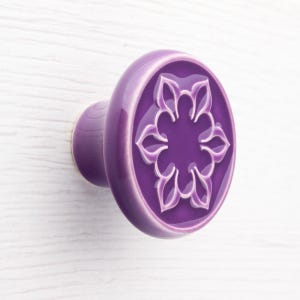 Op de afbeelding: Een paarse keramische kastknop met een bloemmotief. De knop is een levendige paarse tint met een verhoogd, wit bloemmotief in het midden. De knop is gemonteerd op een wit oppervlak.