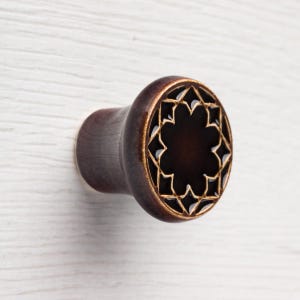 Może przedstawiać: Ciemnobrązowa drewniana zatyczka do ucha z dekoracyjnym wzorem gwiazdy z przodu. Gwiazda jest obrysowana jaśniejszym brązowym kolorem. Zatyczka ma zaokrąglony kształt i gładkie wykończenie.